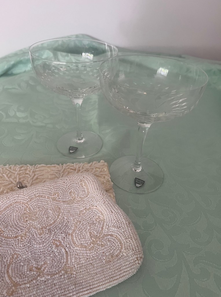 Two crystal champagne glasses on a light blue-green table cloth with two beaded clutch purses in the foreground.