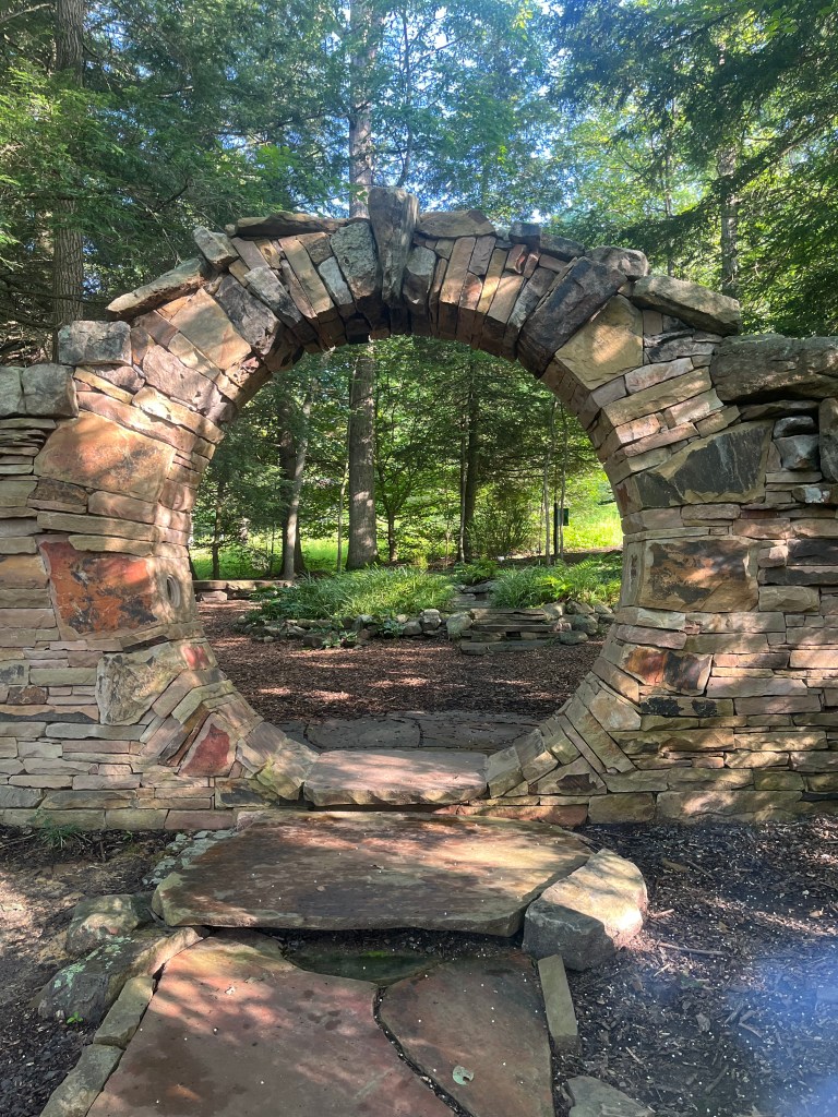 A stone wall with a large circle that allows people to pass through to the other side to a garden and hiking area.
