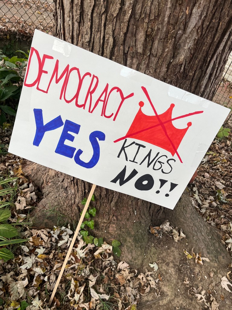 A sign saying "Democracy Yes Kings NO!!" with a red crown with an X through it leans against the trunk of a tree.