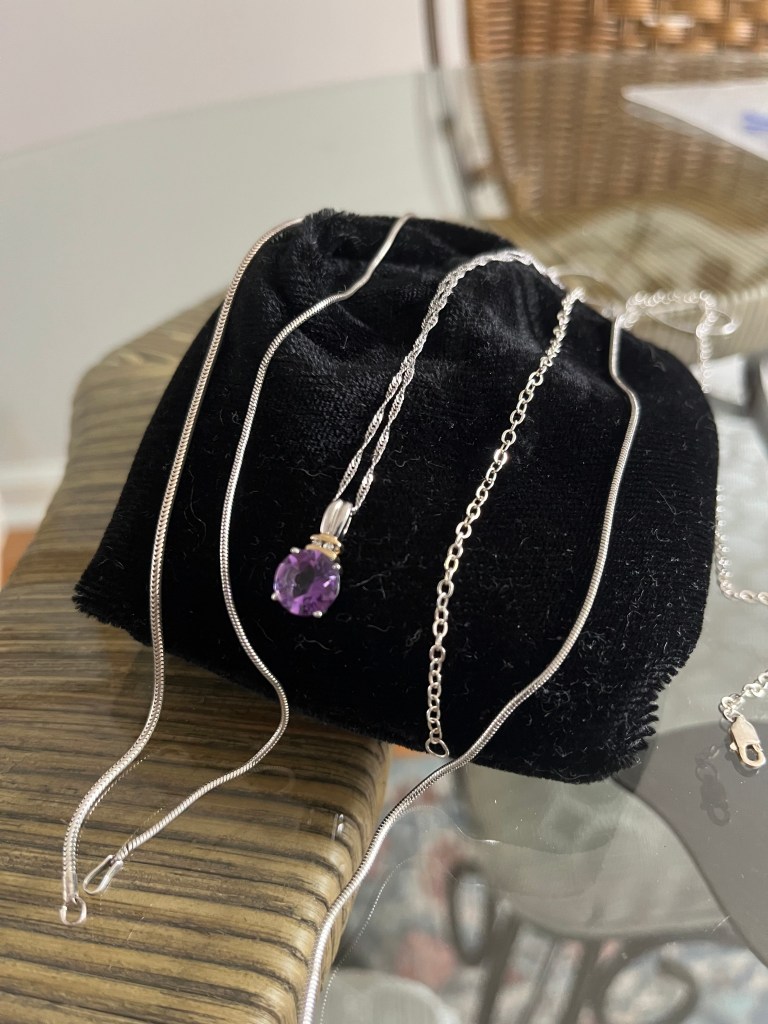 An amethyst pendant on a silver chain lays over a black velvet jewelry bad on a glass table.